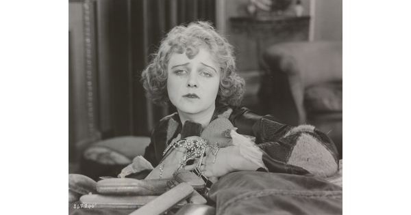A black-and-white photograph of a White woman holding jewelry to her chest as she sadly gazes into the distance.