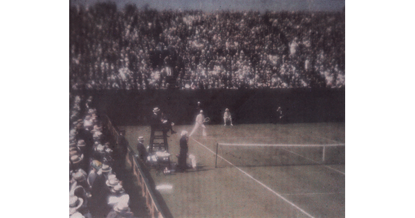 A faded color photograph of spectators in crowded stands watching a tennis match.