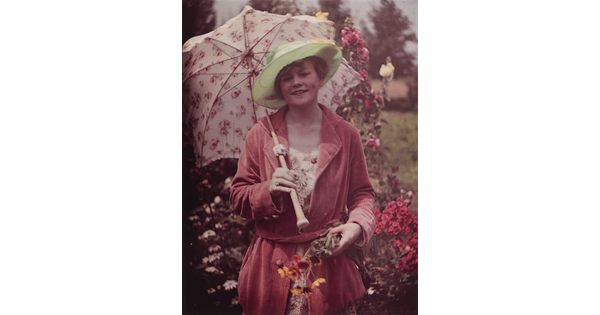 A color photograph of a smiling White woman, standing in a garden, wearing a green wide brim hat, a pink robe, holding a parasol in one hand and a bunch of flowers in the other.