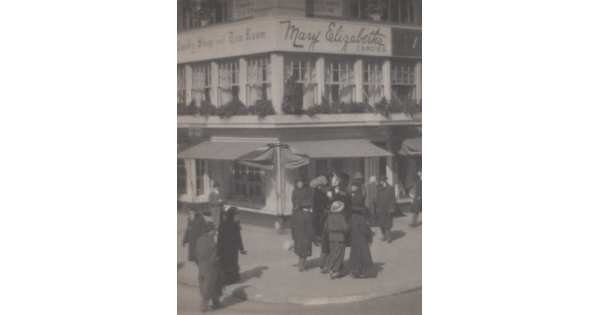 A black-and-white photograph of people walking along a street corner in front of a multi-story building with awnings at street level.