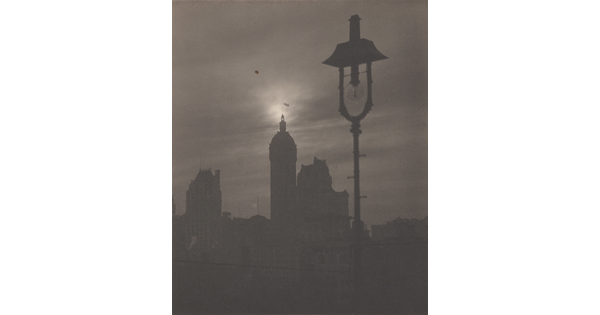 A black-and-white photograph of the silhouettes of several buildings and a gas streetlight.