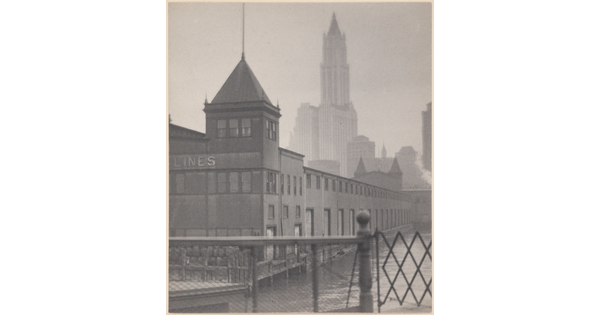 A black-and-white photograph of a low building with a tower on a canal, a gate in the foreground, and skyscrapers in the background.