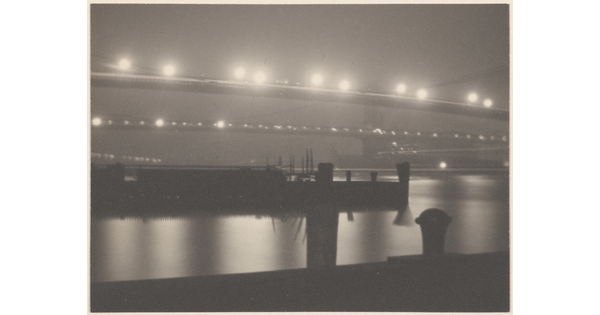 A black-and-white photograph of illuminated bridges at night reflected in the water beneath them; several ships in the water.