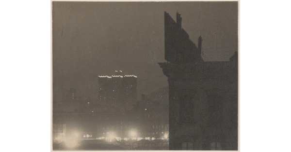 A black-and-white photograph of a building lit around the roofline and a silhouette of a closer building with a billboard on top.