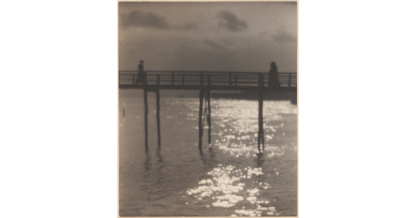 A black-and-white photograph of people walking toward each other on a pier with light reflected over the water underneath.