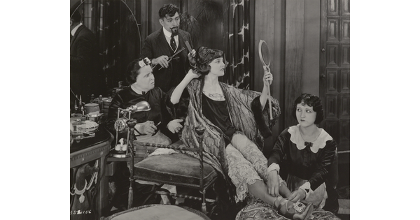 A black-and-white photograph of an elaborately-dressed White woman seated on a bench, gazing into a hand mirror, surrounded by people who are helping her dress.