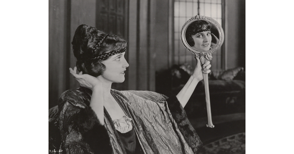 A black-and-white photograph of an elaborately dressed White woman holding up a mirror and gazing into it, her gaze in the mirror meeting the viewer&#039;s eye.
