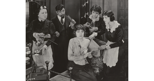 A black-and-white photograph of a White woman seated on a bench surrounded by people who are helping her undress.