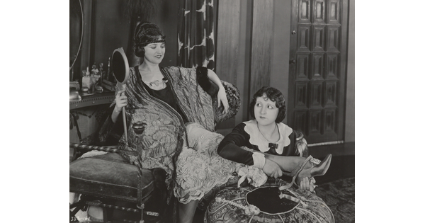 A black-and-white photograph of an elaborately-dressed White woman holding a large hand mirror, seated on a chair with one foot resting on an ottoman as a maid puts on her shoes.