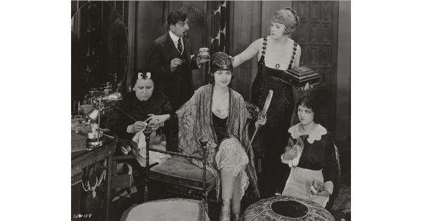 A black-and-white photograph of an elaborately-dressed White woman seated on a bench surrounded by people who are working on her hair and nails.
