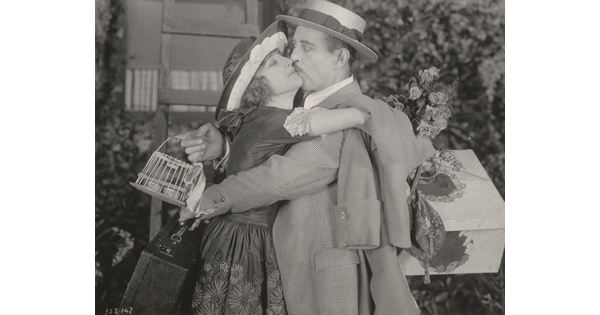 A black-and-white photograph of a White woman eagerly embracing and kissing a White man who is taken by surprise.