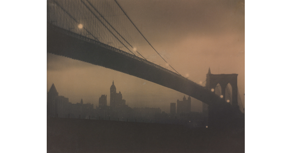 A sepia-toned photograph of a long bridge with a foggy cityscape in the background taken at night.