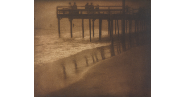 A sepia-toned photograph of people standing on a pier as waves crash beneath it.