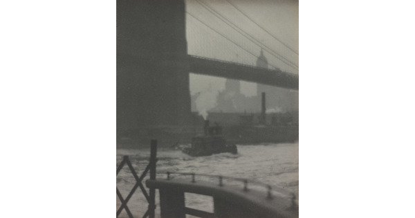 A black-and-white photograph of a suspension bridge seen from below, a tugboat on the water, and a city skyline in the distance.