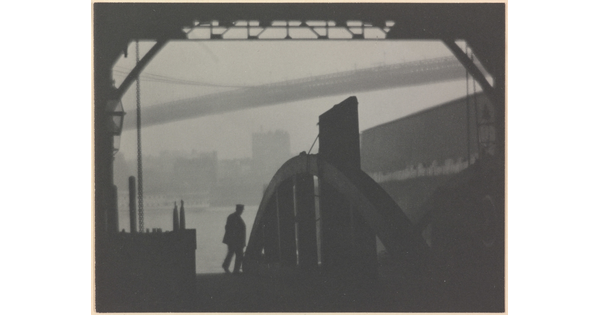 A black-and-white photograph of the silhouette of a person walking on a dock with a hazy city and a large bridge in the background.