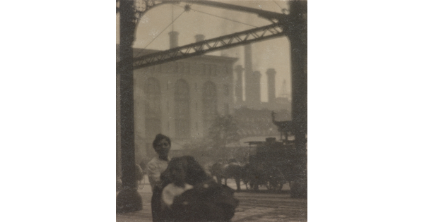 A black-and-white photograph of people and horse-drawn carriages on a street in front of a factory.