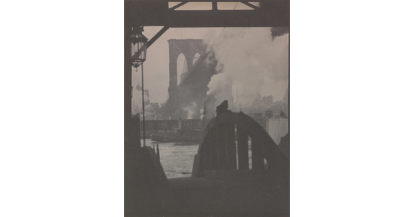 A black-and-white photograph of a dock in silhouette with a hazy city and a large bridge in the background.