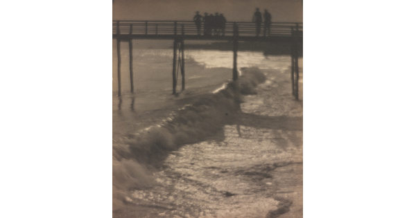 A sepia-toned photograph of people standing on a pier as waves crash beneath it.