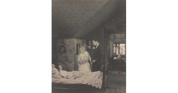 A black-and-white photograph of a White woman lying in bed as an elderly woman and a tall young man watch over her.