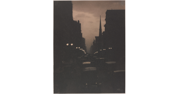 A sepia-toned photograph of a busy city street at dusk illuminated by street lamps and car lights.