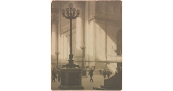 A sepia-toned photograph of people walking through a sunlit plaza with towering light posts and columns.
