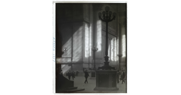 A black-and-white photograph of people walking through a sunlit plaza with towering light posts and columns.