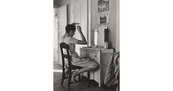 A black-and-white photograph of a White teenage girl seated in front of a mirror on top of a dresser fixing her hair.