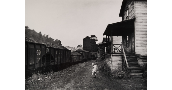 A black-and-white photograph of a young girl carrying a pail between a train and some clapboard buildings.