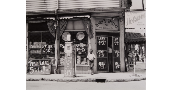 A black-and-white photograph of a dark-skinned boy standing in front of a grocery store.