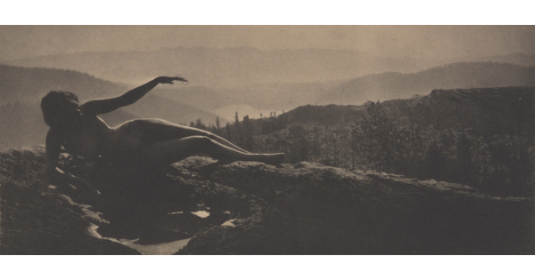 A sepia-toned photograph of a nude woman in shadow lying on a hilltop with cascading hills behind her.