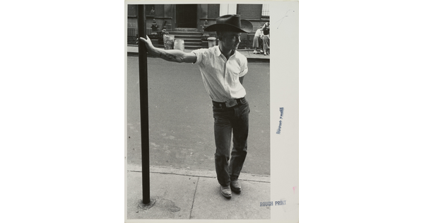 A black-and-white photograph of a White man in a cowboy hat leaning against a metal pole on a city sidewalk.