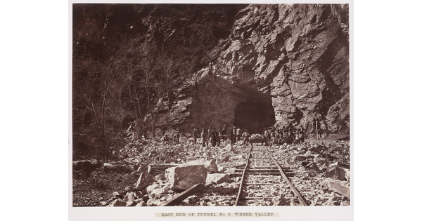 A black-and-white photograph of railroad tracks leading into a rocky tunnel, with workers gathered at the  tunnel entry.