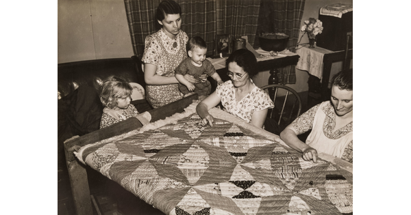A black-and-white photograph of two seated White women quilting, a woman with a child on her lap, and another child seated next to her.