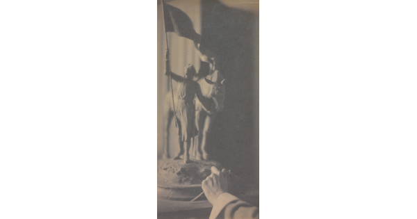 A vertically-oriented black-and-white photograph of a hand reaching toward a sculpture of a girl holding a flag in one hand and a horse's rein in the other.