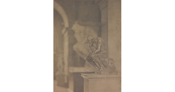 A black-and-white photograph of a small sculpture of a sitting man resting his head on his fist and elbow on one knee in front of a larger sculpture of the same.