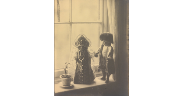 A sepia-toned photograph of two light-colored dolls in Russian imperial clothing standing next to a flower pot on a window sill.