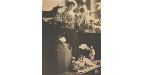 A sepia-toned photograph of several White women working marionettes above a puppet stage.