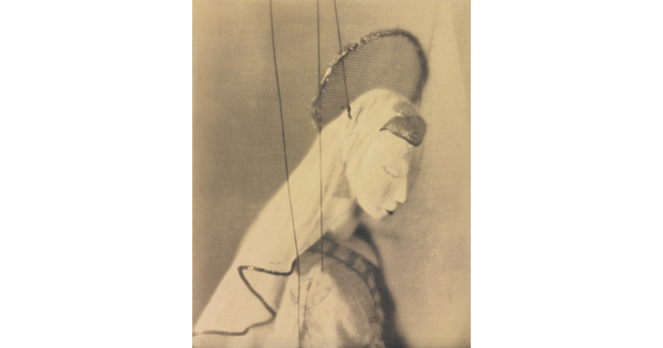 A sepia-toned close-up photograph of the head and shoulders of a marionette with a halo and veil.