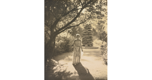 A black-and-white photograph of a light-skinned woman in a flowing dress walking among trees with the sun at her back.