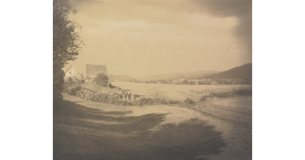 A sepia-toned photograph of an open field with a house and a barn in the distance.