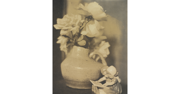 A sepia-toned photograph of roses in a vase and a figurine of two young children reading a book.
