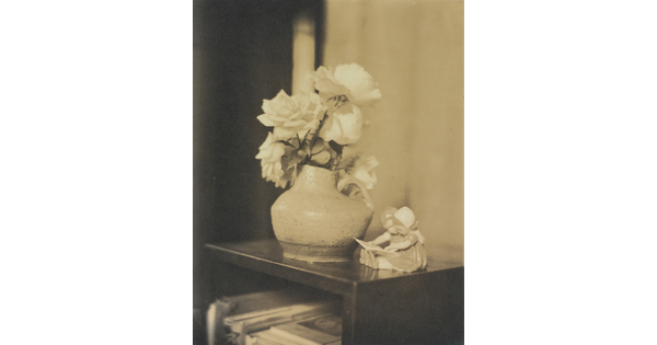 A sepia-toned photograph of roses in a vase and a figurine of two young children reading a book on a shelf.