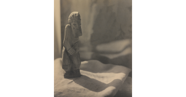 A sepia-toned photograph of a rough-hewn sculpture of a bearded man standing on a piece of folded fabric.