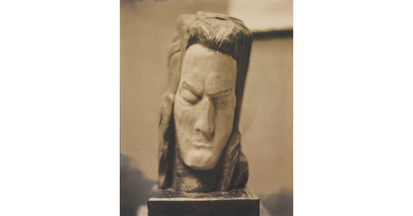 A sepia-toned photograph of a rough-hewn sculpture of a man's head resting on a pedestal.