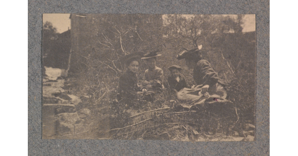 A black-and-white photograph of a group of four light-skinned women wearing hats and dresses sitting among trees and bushes.