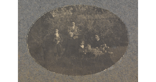 An oval-shaped black-and-white photograph of a group of light-skinned women and children holding flower bouquets in a natural setting.
