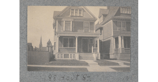 A black-and-white photograph of a three-story wood house, with a front porch and steps leading to a sidewalk in front.