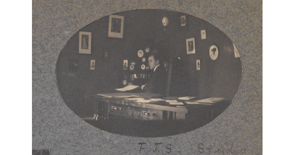 An oval-shaped black-and-white photograph of a White man seated behind a desk; framed pictures hang on the walls behind him.