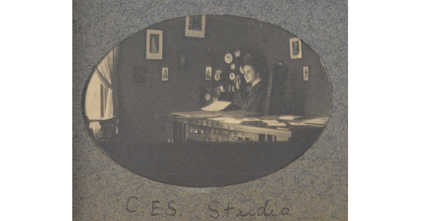 An oval-shaped black-and-white photograph of a White woman seated at a desk; framed pictures hang on the walls behind her.