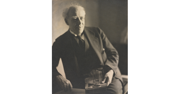 A black-and-white photograph of an older White man with white hair, wearing a dark suit and tie, leaning on one arm while holding an object in his hand.